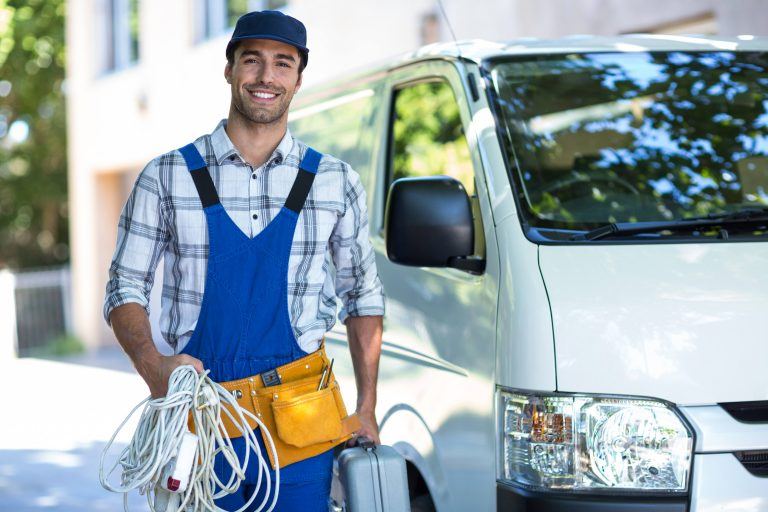 Smiling technician in overalls standing next to a white van, holding tools and electrical cords.